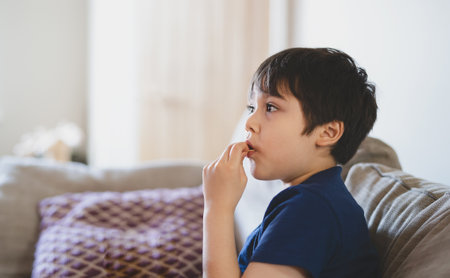 Portrait New normal lifestyle Kid siting on sofa eating potato crisps while watching TV, Young boy eating snack while watching cartoon, Child stay at home during social distance from Covid-19の写真素材