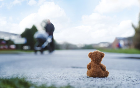 Rear view Lonely Teddy bear doll sitting alone on hailstones with blurry father pushing stroller walking on footpath.lost brown bear toy looking out on the road, International missing children's dayの写真素材