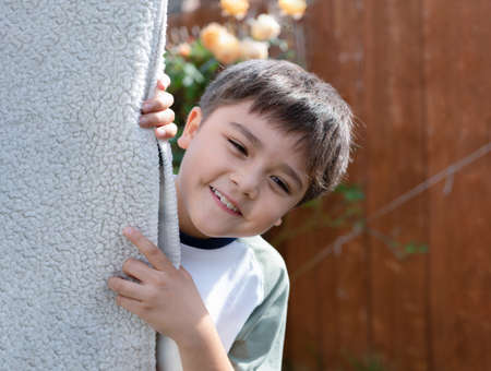 Happy kid playing hide and seek in the garden, Cute boy with smiling face while standing behind blanket hanging on clothesline, Funny child playing outdoor on sunny day summer.の写真素材