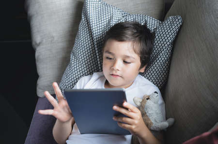 Shool kid doing online home work on tablet, Happy boy enjoy playing game on digital pad, Kid hugging dog toy lying down on sofa while watching or reading reading story on ebookの写真素材