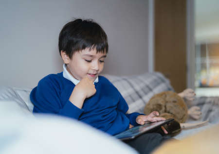 Happy schoolboy with smiling face playing game online with friend on tablet,Kid using internet sending homework to the teacher,Positive child sitting on sofa relaxing alone in living room after Schoolの写真素材