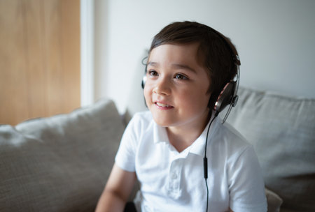 Close up kid face wearing headphone having fun playing game online with friends, Cute School boy sitting on sofa looking up watching TV with smiling face, Young boy with headset listening to musicの写真素材