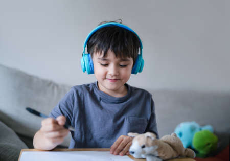 Happy kid wearing headphones listening to music while drawing on paper, Indoors portrait Cute child boy enjoy creative activity at home on weekend.の写真素材