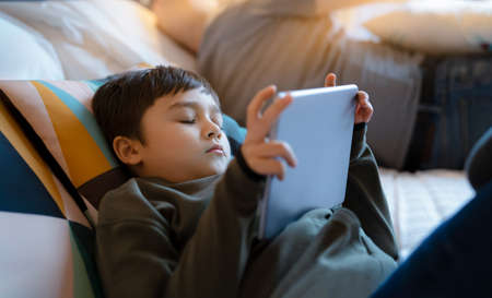 Young boy falling sleep while watching cartoon on tablet, Tried kid lying in bed play game online,...