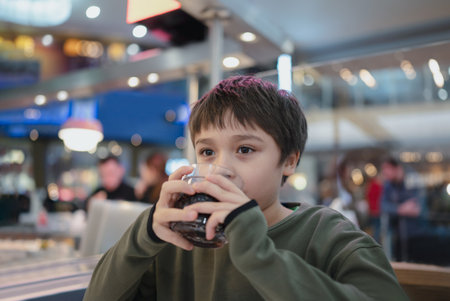 Portrait of  Young kid sitting on table drinking cold drink in restaurant, A boy drinking soda or soft drink from glass, Child boy waiting for food in cafeの写真素材