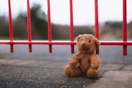 Lost teddy bear toy sitting next to metal fence at playground in gloomy day, Lonely and sad brown bear doll lied down alone in the park, lost toy or Loneliness concept, International missing Children dayの写真素材