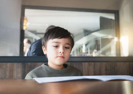 A School kid reading or choosing food in the menu ready to order food for lunch, Child boy sitting and waiting for his meal in the restaurant.の写真素材