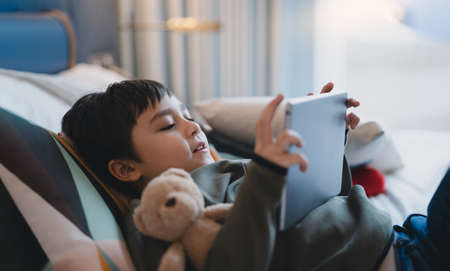 Happy Kid lying in bed holding tablet watching cartoon and chatting with friends on digital pad, Cute young boy playing games online on internet, Child relaxing at home with familyの写真素材
