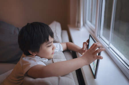 Cinematic portrait a happy young boy playing a game online on the tablet, Kid watching cartoon from internet, Child sitting on sofa next to window with a raining day in the morningの写真素材