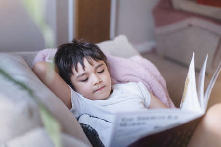 Happy School kid lying on sofa reading a book with morning light shining from window. Child boy...
