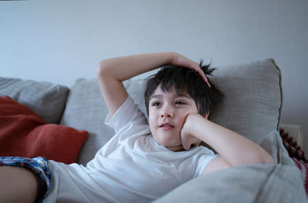 Portrait young boy with curious face sitting on sofa watching cartoon, School Kid sitting alone on couch relaxing in living room after back from school.の写真素材
