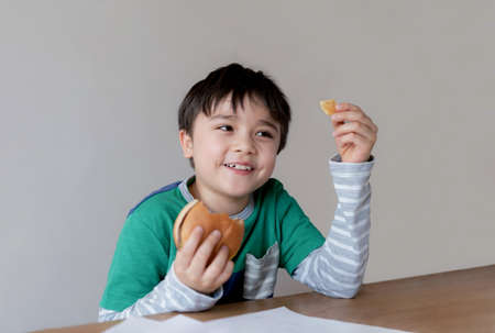 Healthy young boy eating burger. A child holding cheeseburger and looking at chips on his hand with smiling face, Happy Kid having fast food hamburger for snack. Favorite children's food.の写真素材