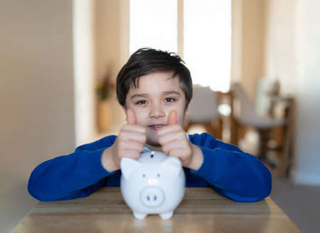 Happy School boy putting thumbs up on piggy bank and looking at camera with smiling face.Child showing money saving box.kid Learning financial responsibility and planning about saving money for futureの写真素材
