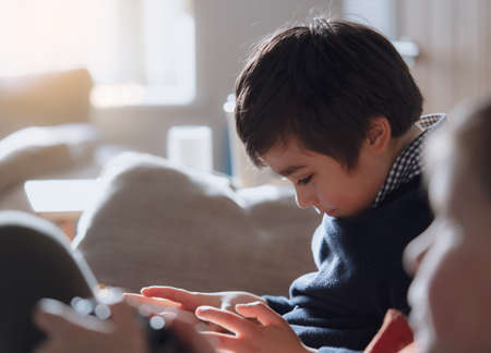 Cinematic portrait young boy playing game on tablet sitting on couch with sunlight shining from...