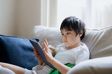 Young boy using tablet playing game on internet, Kid sitting on sofa watching or talking with friend...