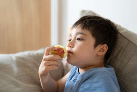 Healthy child eating red apple, Cute boy eating fresh fruit for his snack while watching TV in living room, Close up kid face eating food. Healthy food for children conceptの写真素材