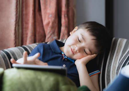 Cinematic portrait young boy playing game on tablet sitting on sofa with light shining from window, Kid playing games online on internet at home, Child talking video call with friends at homeの写真素材