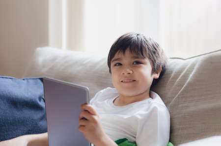 Cinematic portrait young boy playing game on tablet sitting on sofa with light shining from window,...