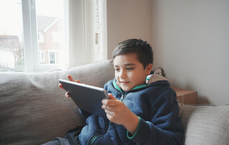 Education concept,School Kid holding tablet reading E-book for homework,Portrait happy Child playing game online on internet with friends,Young boy watching cartoon on digital pad,Kid sitting on sofaの写真素材