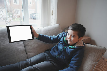 Happy Child with tablet computer. Portrait Kid Holding And Showing Digital Tablet With Empty Screen  Mockup, Young boy sitting on sofa relaxing and playing with touch pad at homeの写真素材