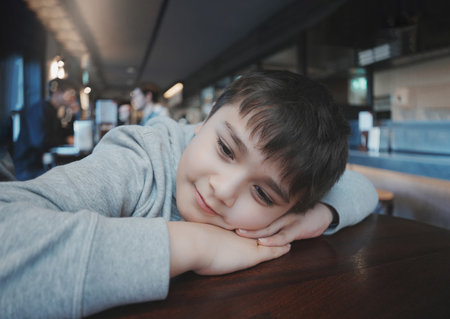 Indoors portrait Young boy with smiling face sitting alone in coffee shop, cafe waiting for food, Cinematic Portrait Kid looking deep in thought,Child looking out with thinking face.Healthy lifestyleの写真素材
