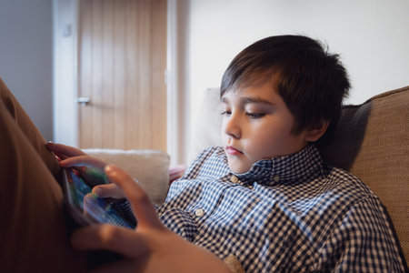 Education concept,School Kid holding tablet reading E-book for homework,Portrait happy Child playing game online on internet with friends,Young boy watching cartoon on digital pad,Kid sitting on sofaの写真素材