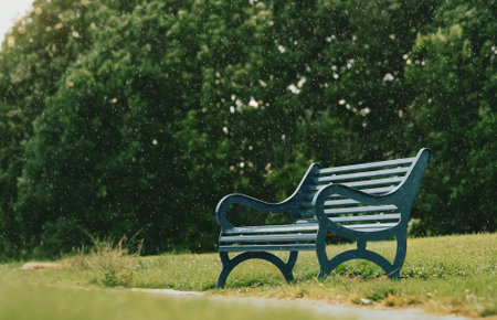 Bench in the local park with blurry rain falling on Spring forest background, Blue Metal stylish Bench in the Park with Green Lawn on Summer Background, Retro vintage toneの写真素材