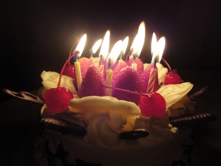 birthday cake with sweet cherry, strawberry,white rose chocolate and light candle in the darkの写真素材