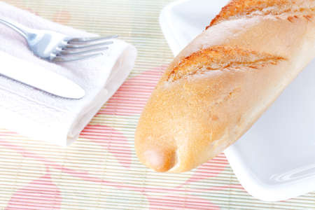 Crisp fresh-baked baguette on white plate, napkin, fork and knife on bamboo placemat