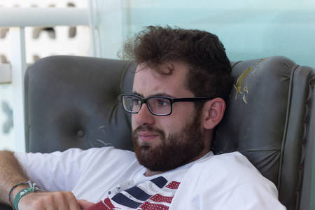 Young caucasian guy with beard and glasses sitting on old vintage sofa on light colored wall...