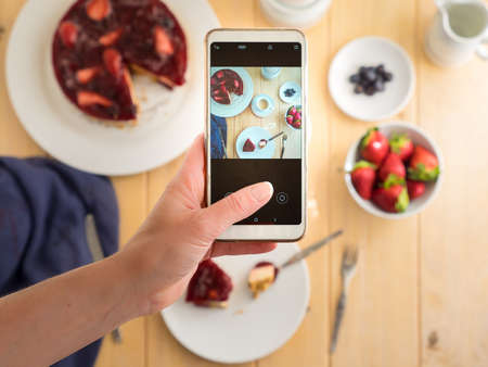 Hand with mobile phone taking a picture of a cheesecake with red fruits. Photo on phone. Selective focus. Concept home cooking.の写真素材