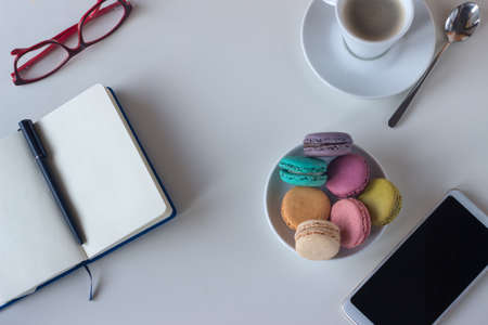 Work table with writing pad, glasses, coffee cup, mobile, and plate full of colorful macaroons, on light background. Copy spaceの写真素材