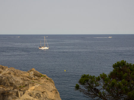 Small sailboat seen from the rocks, sailing on a silver sea on a sunny day. Vacation concept.の写真素材
