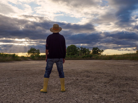 With rubber boots for the dry field. The farmer goes with his rubber boots and his shovel to a barren field. See from behind. Drought concept.の写真素材
