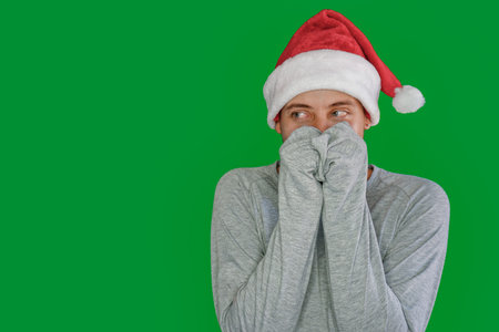Young Latino man in Christmas hat looking to the side with his hands over his mouth and gray sweatshirt.の写真素材