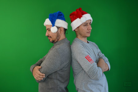 two Latino men in blue and red Christmas hats turn their backs to each other as they cross their arms and stare negatively at each other. Christmas concept.の写真素材