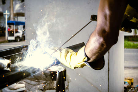 worker person hands with yellow gloves welding metal in workshop with blue fire and sparks