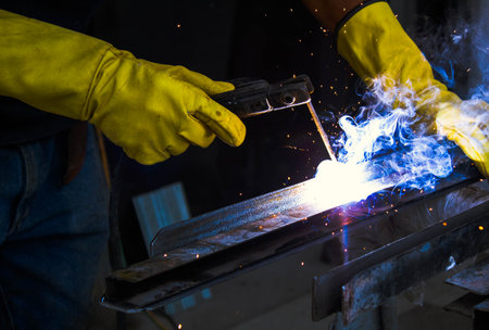 worker person hands with yellow gloves welding metal in workshop with blue fire and sparks