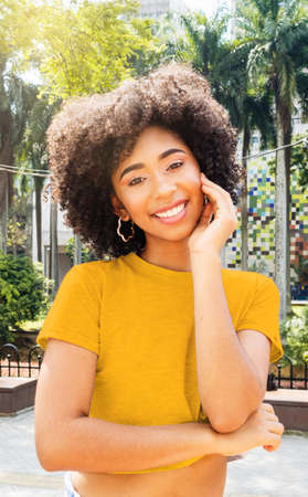 portrait of beautiful afro woman in yellow shirt in public park outdoors smiling at camera on sunny dayの写真素材