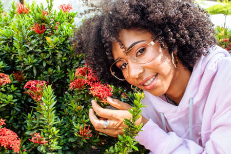 beautiful african american girl touching garden flowers looking at camera wearing glasses and sweatshirt outdoors on sunny dayの写真素材