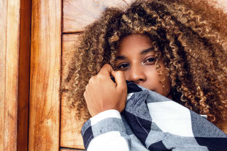 portrait of curly haired afro young teen woman in blue jacket covering her face in fashion on wooden background.の写真素材
