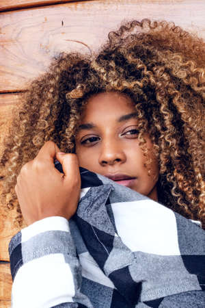 portrait of curly haired afro young teen woman in blue jacket covering her face in fashion on wooden...
