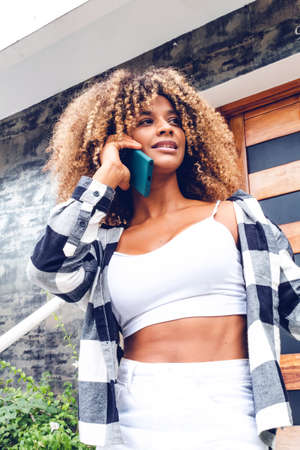 beautiful afro curly hair woman wearing sweater talking on the phone at the door of her house cheerful happy.の写真素材