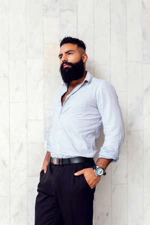 Caucasian handsome man with fashionable stylish long beard, male business person posing at white wall of building outdoorsの写真素材