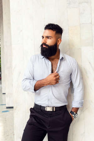 Caucasian handsome man with fashionable stylish long beard, male business person posing at white wall of building outdoorsの写真素材
