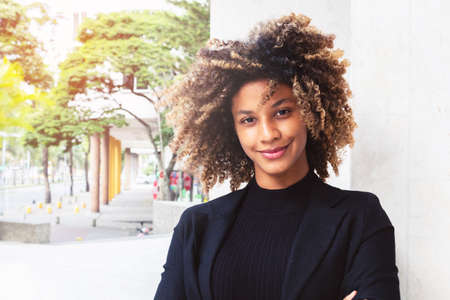 portrait of beautiful adult business woman, wearing elegant black suit looking at camera smiling happy in building on sunny day, afro woman with curly hairの写真素材