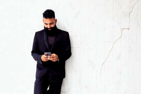 Caucasian adult man with elegant long beard wearing black suit using mobile phone outdoors for business smiling leaning on isolated white wall.の写真素材