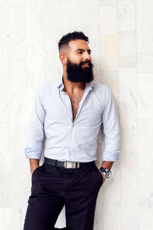 Caucasian handsome man with fashionable stylish long beard, male business person posing at white wall of building outdoorsの写真素材