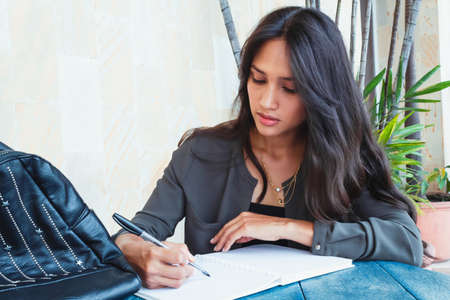 Latin teen student girl outdoors writing in her notebook sitting at the table smiling, doing her...