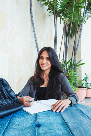 Latin teen student girl outdoors writing in her notebook sitting at the table smiling looking at camera, doing her homework. going back to class.の写真素材